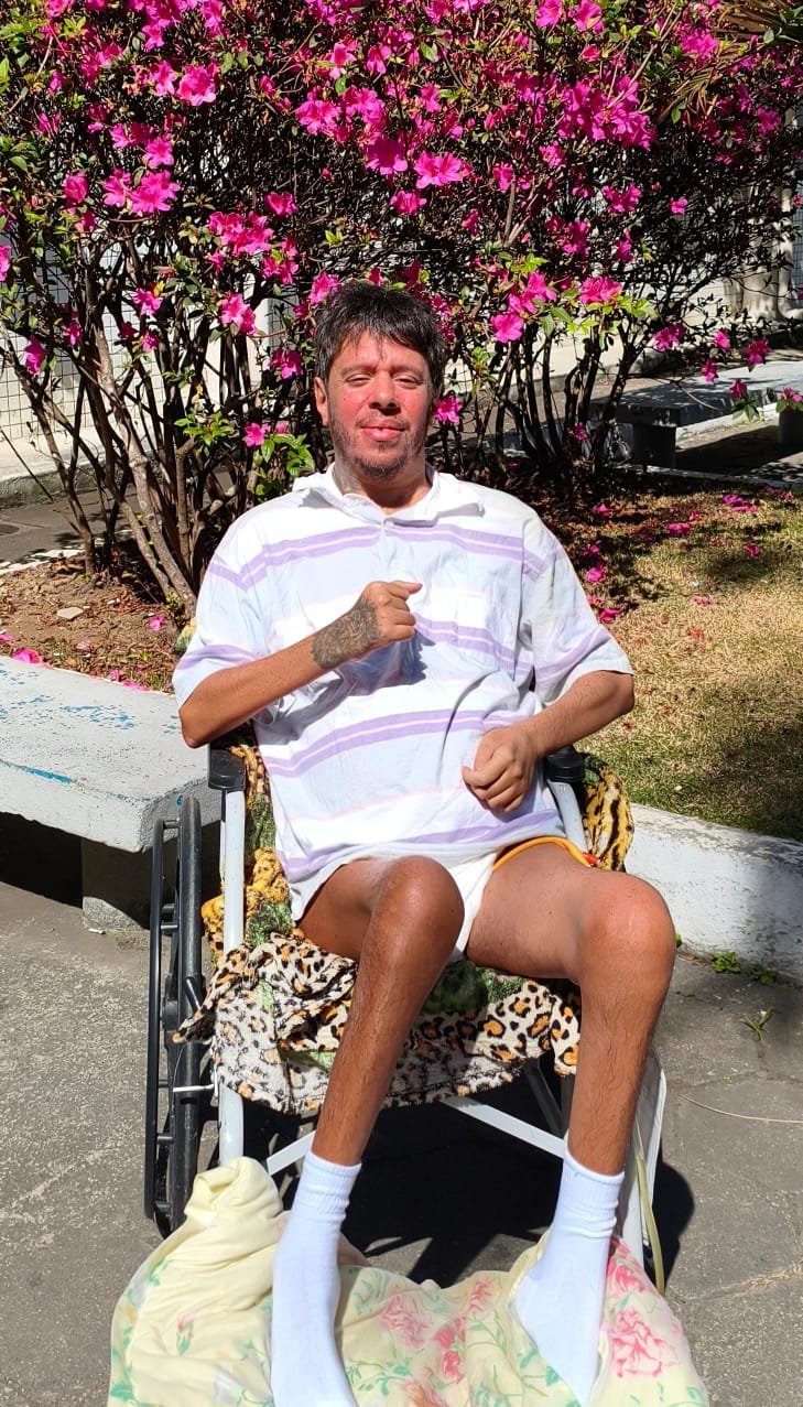 Berguim sorrindo ao ar livre, sentado em sua cadeira de rodas, em frente a uma azaleia florida.